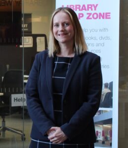 A photo of a woman smiling in front of an academic library with a library poster on the window behind her